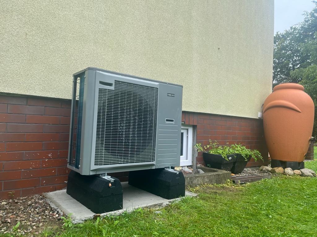 Szara jednostka zewnętrzna klimatyzatora zamontowana na betonowej podstawie i czarnych podkładkach, ustawiona przy ceglanej ścianie budynku z jasną elewacją i dużą, glinianą amforą w tle.