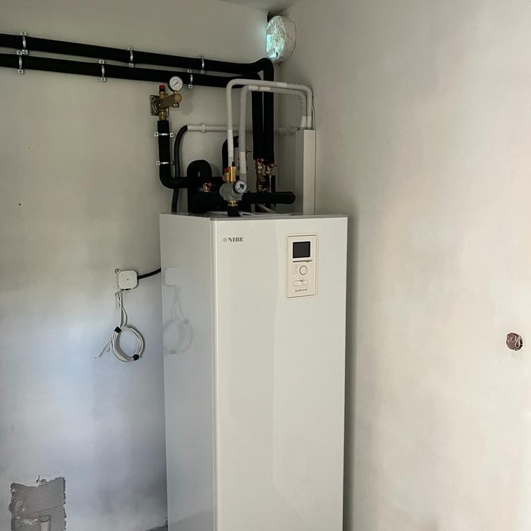 Biała pompa ciepła Nibe zainstalowana w pomieszczeniu z widocznymi rurami i elementami instalacji, niedokończone wykończenie ściany.