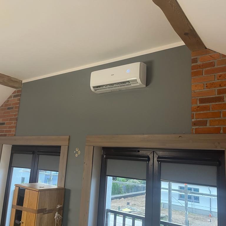 Biała jednostka klimatyzacji zamontowana na szarej ścianie w pokoju z ceglanymi i drewnianymi elementami dekoracyjnymi, widok z dołu na okna z roletami.