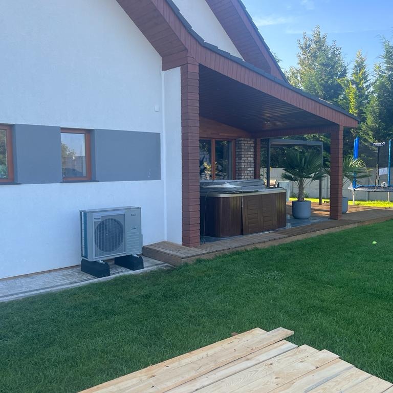 Klimatyzator zewnętrzny zamontowany na elewacji domu jednorodzinnego, obok tarasu z jacuzzi i zielonego trawnika, na pierwszym planie deski.