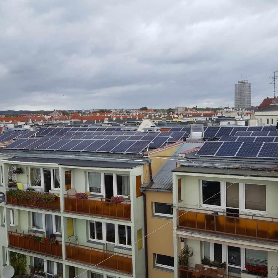 Widok z góry na dachy budynków mieszkalnych z zainstalowanymi panelami słonecznymi, w tle panorama miasta pod pochmurnym niebem.