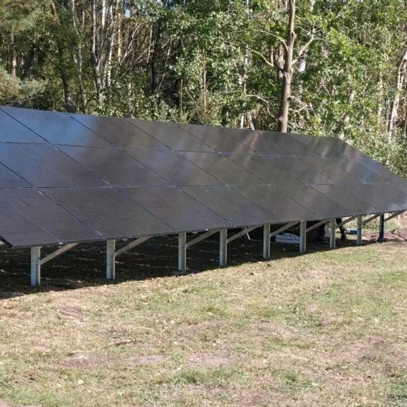 Rząd paneli słonecznych na metalowej konstrukcji, ustawionych na trawniku przed lasem, widoczne połączenia modułów.