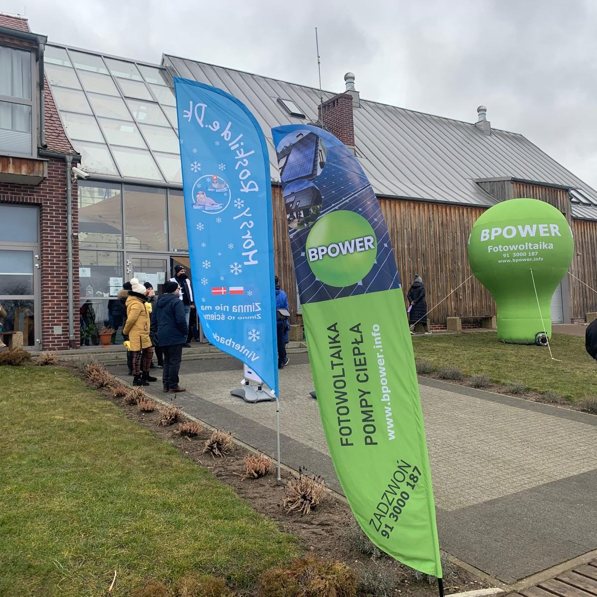 Zewnętrzny widok budynku z dwoma flagami reklamowymi i dużą zieloną dmuchaną żarówką z logo BPOWER, promującymi fotowoltaikę i pompy ciepła.