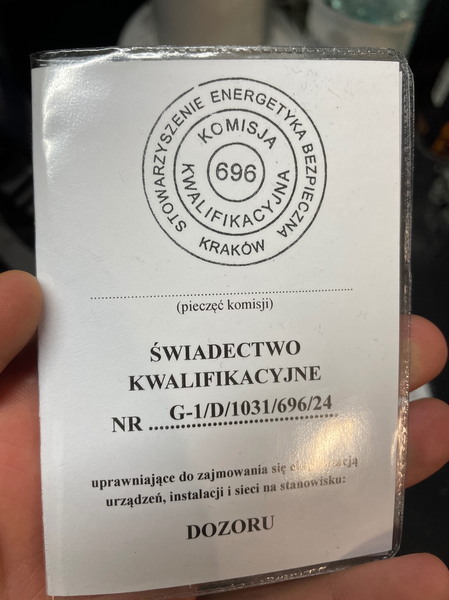Zdjęcie świadectwa kwalifikacyjnego elektryka dozoru, wydanego przez Stowarzyszenie Energetyka Bezpieczna w Krakowie, numer G-1/D/1031/696/24, trzymanego w dłoni.