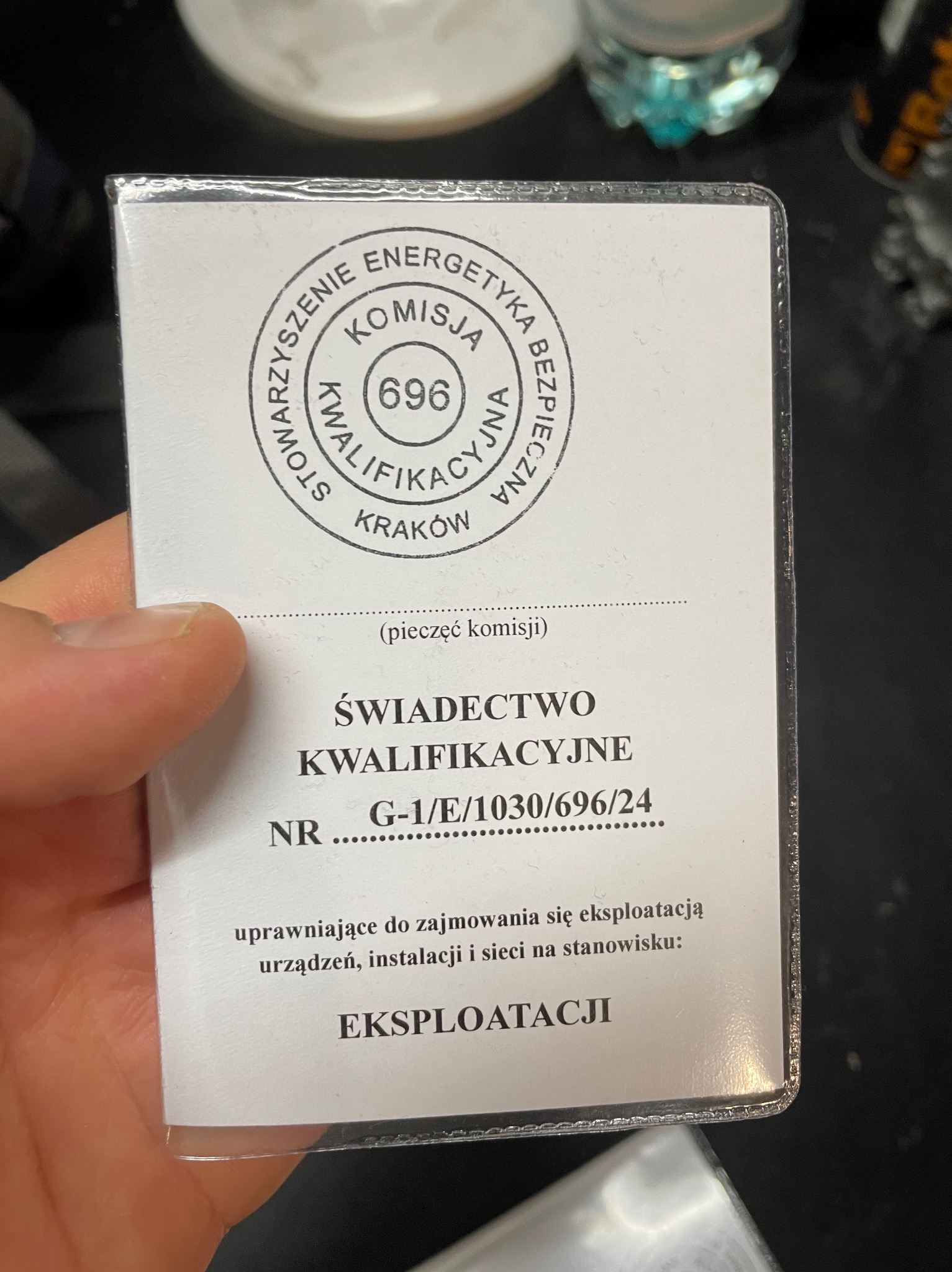 Świadectwo Kwalifikacyjne G-1/E/1030/696/24, uprawniające do zajmowania się eksploatacją urządzeń, instalacji i sieci, trzymane w dłoni na ciemnym tle.