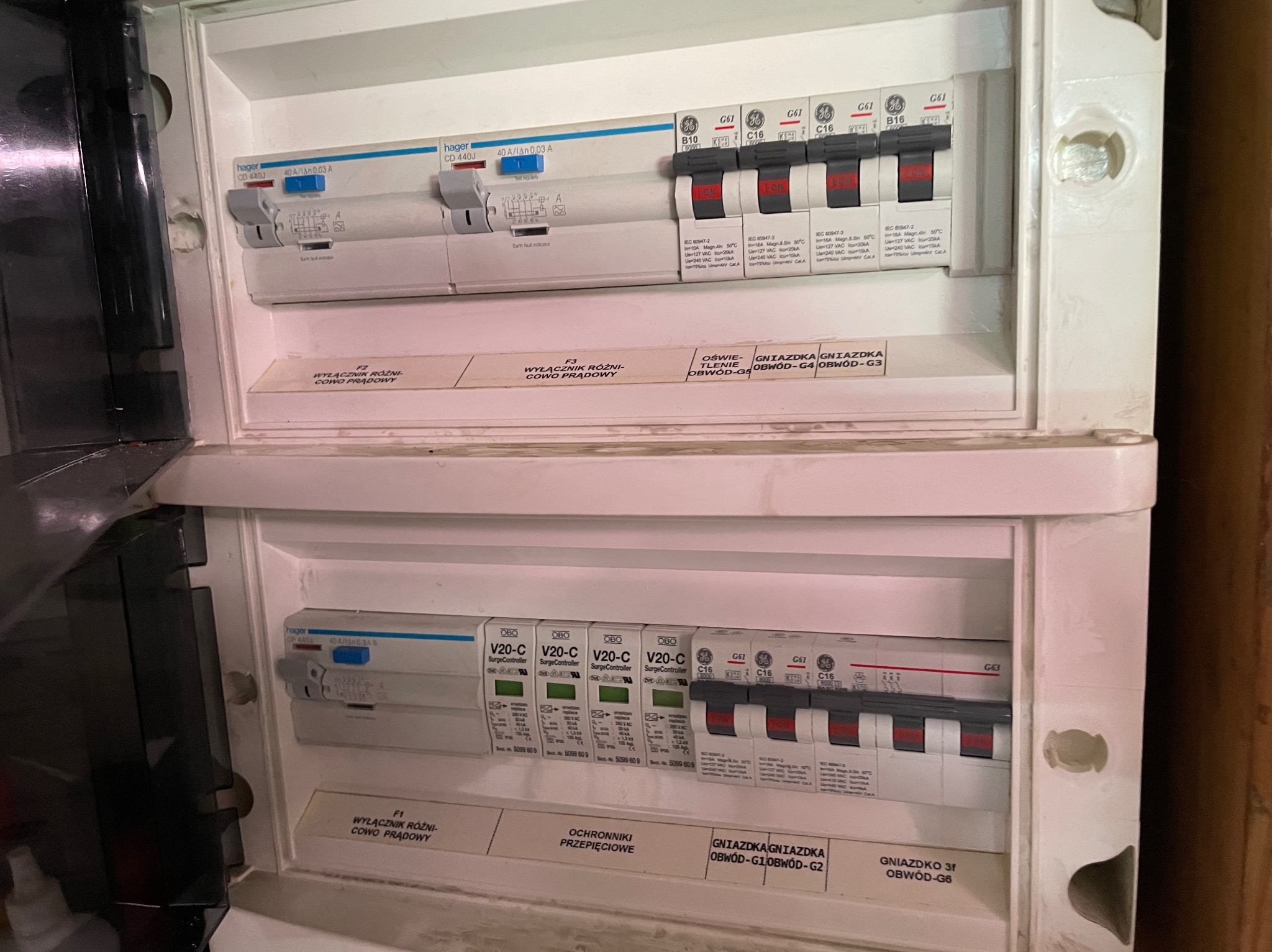 Rozdzielnica elektryczna z dwoma rzędami zabezpieczeń: wyłączniki różnicowoprądowe Hager, ograniczniki przepięć OBO V20-C oraz wyłączniki nadprądowe GE, etykiety opisujące obwody.