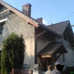 Dom w trakcie budowy z widoczną konstrukcją dachu i kominem z cegły, fragmentem rynny, zaroślami i fragmentem ogrodzenia w kadrze.