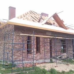 Budynek w trakcie budowy, widoczna murowana elewacja z cegły, rusztowania, drewniana konstrukcja dachu w trakcie montażu, komin z cegły.