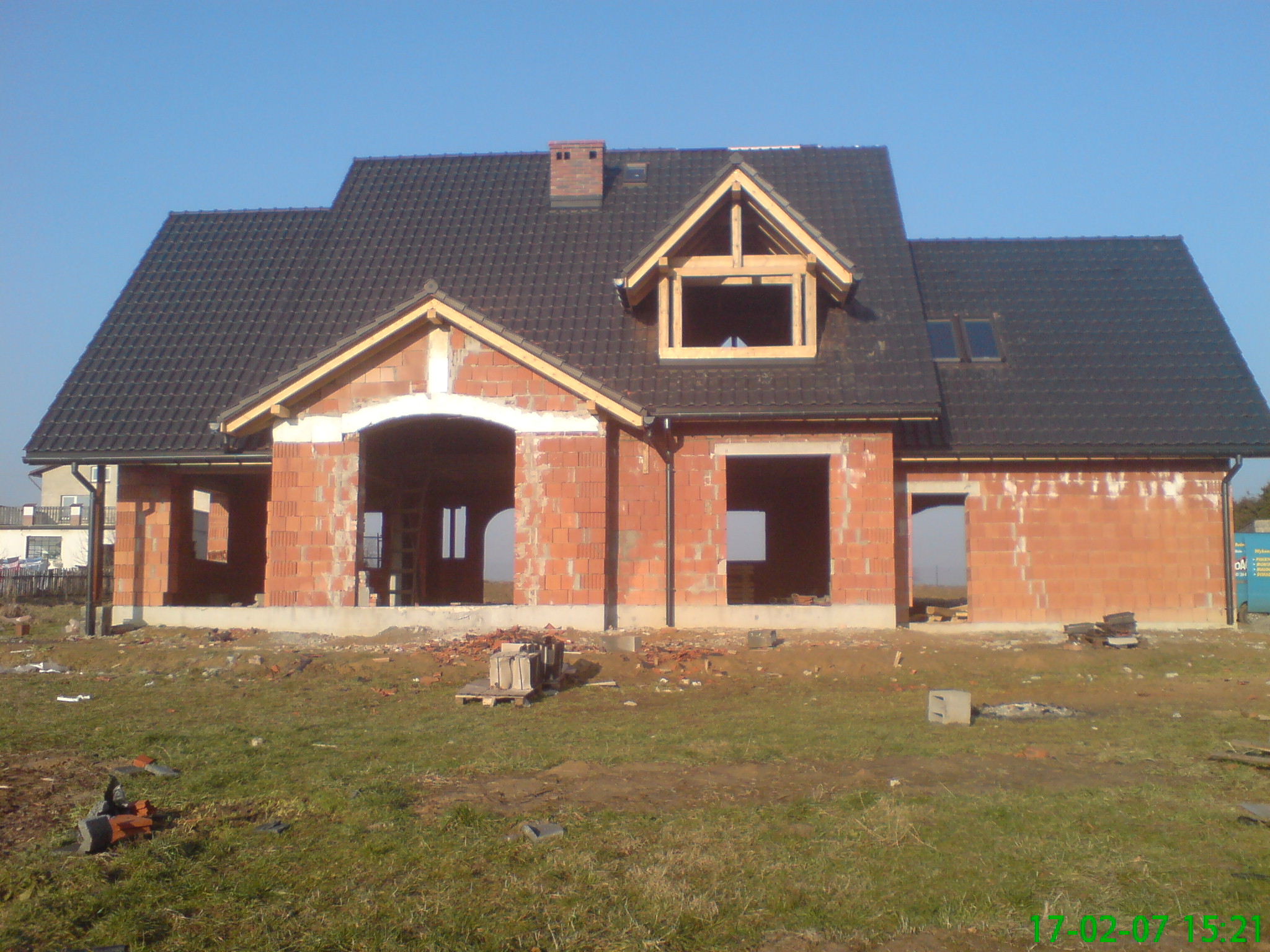 Surowy budynek jednorodzinny z ciemną dachówką, widoczne fragmenty drewnianej konstrukcji dachu i otwory okienne bez ram, na pierwszym planie trawnik z gruzem.