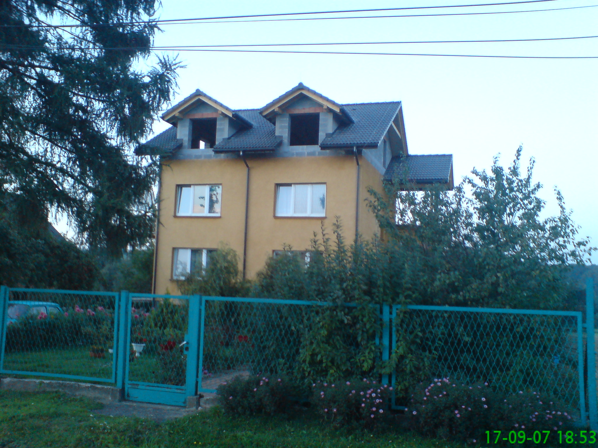 Dwupiętrowy dom w trakcie budowy, z częściowo ukończonym dachem pokrytym szarą dachówką, widoczny przez turkusowe ogrodzenie z siatki.