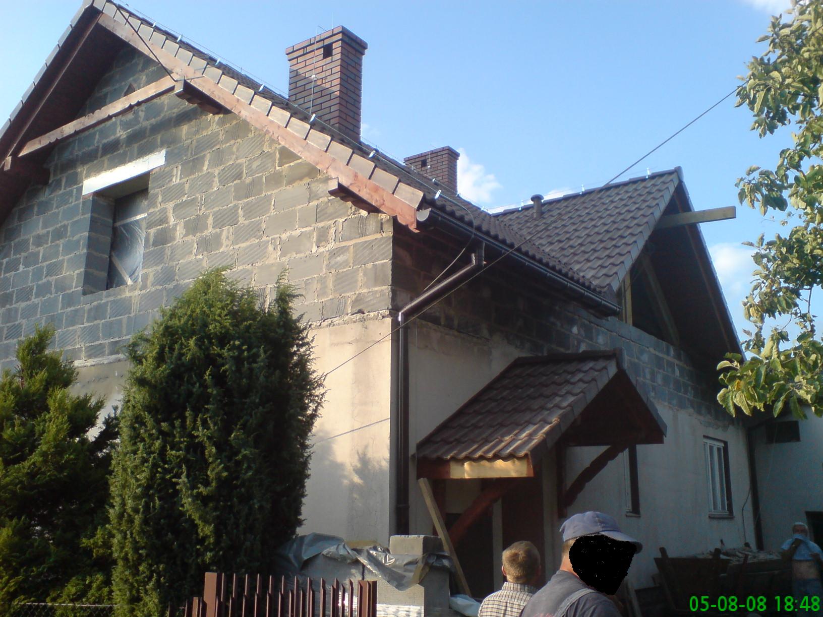 Dom w trakcie budowy z widoczną konstrukcją dachu i kominem z cegły, fragmentem rynny, zaroślami i fragmentem ogrodzenia w kadrze.