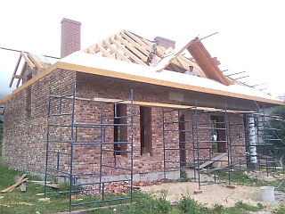 Budynek w trakcie budowy, widoczna murowana elewacja z cegły, rusztowania, drewniana konstrukcja dachu w trakcie montażu, komin z cegły.