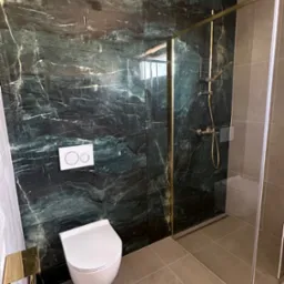 Nowoczesna łazienka z kabiną prysznicową w złotych ramach i toaletą podwieszaną na tle ściany z zielonego marmuru. Widoczne szare płytki podłogowe.