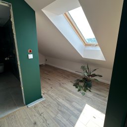 ROMAN MATUSHCHAK ARMBUILDING - Poddasze po remoncie: zielona ściana z gniazdkiem, drewniana podłoga, okno dachowe z widokiem na zieleń, rośliny doniczkowe. Widoczne wejście do łazienki.