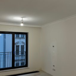 ROMAN MATUSHCHAK ARMBUILDING - Jasne wnętrze w trakcie prac wykończeniowych: białe ściany z listwami, okno balkonowe z widokiem na budynek, fragmenty paneli podłogowych na podłodze.