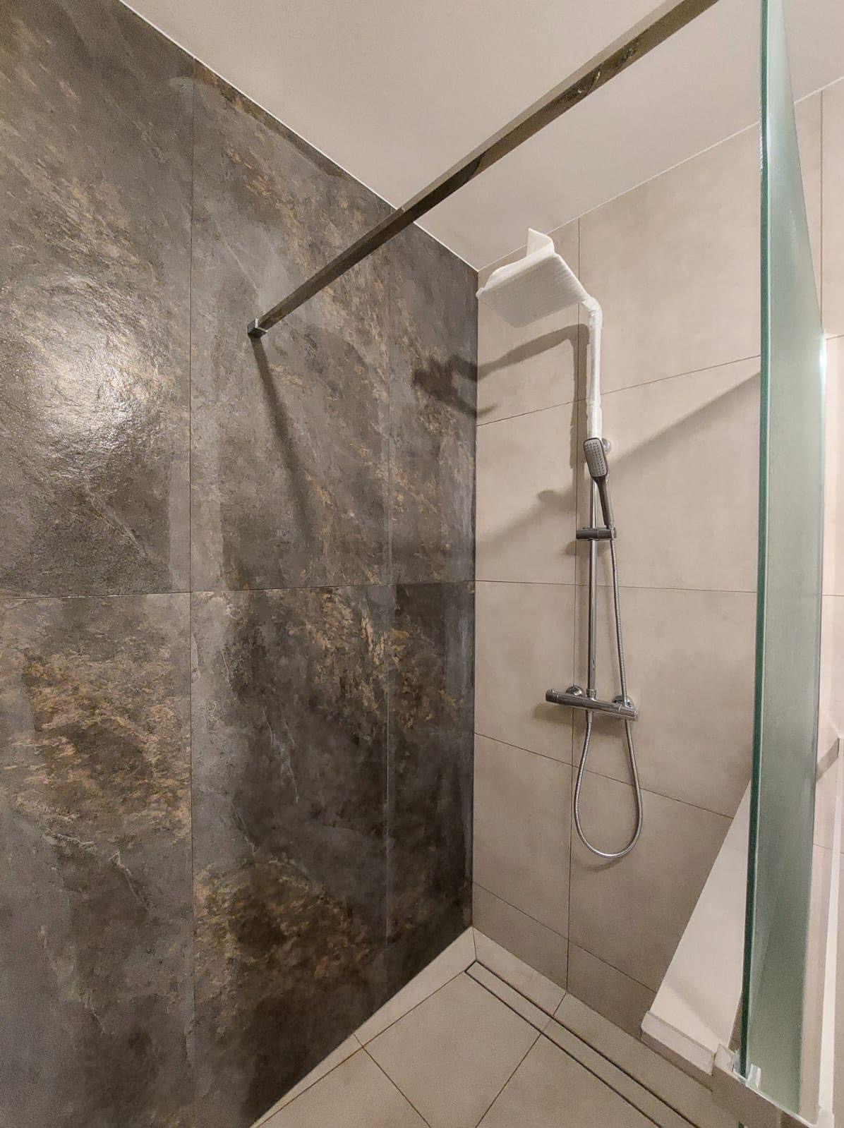 Nowoczesna kabina prysznicowa z deszczownicą, wykończona płytkami w odcieniach szarości i beżu, zamknięta szklaną ścianką. Minimalistyczny design z widocznym odpływem liniowym.