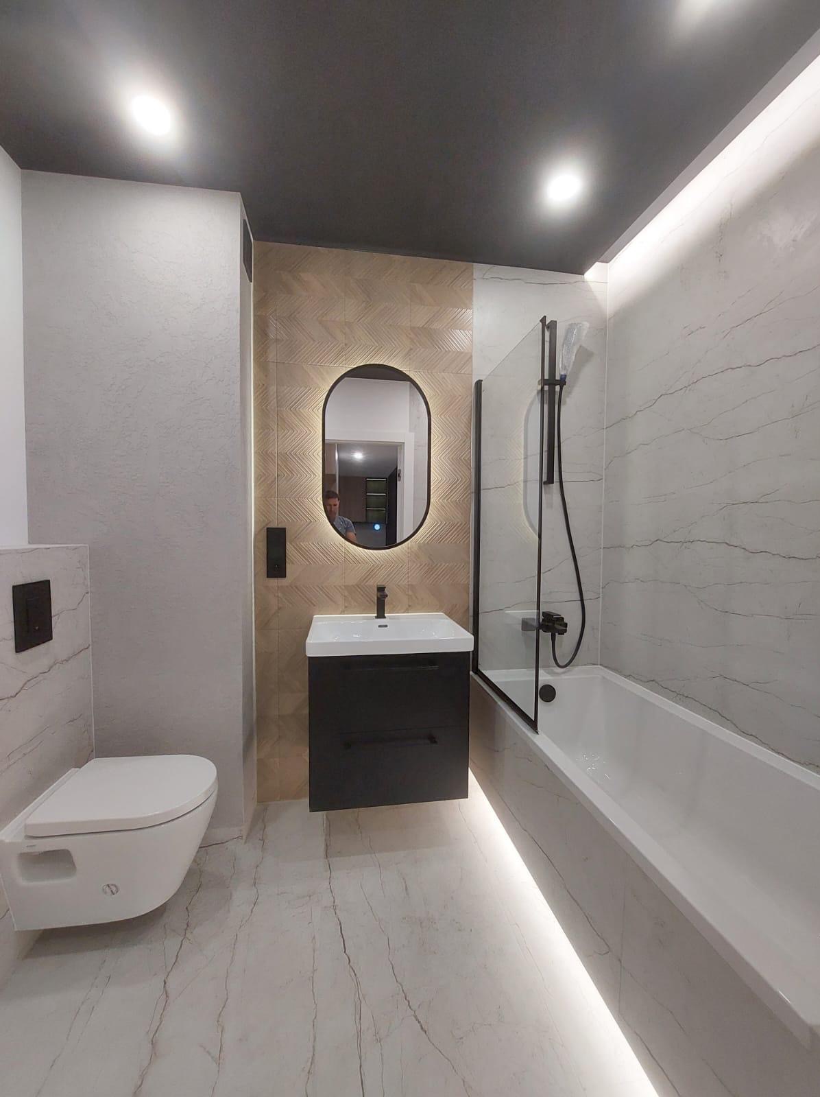 Nowoczesna łazienka z owalnym lustrem z podświetleniem, białą armaturą i marmurowymi płytkami. Czarna szafka pod umywalką i szklany panel prysznicowy.