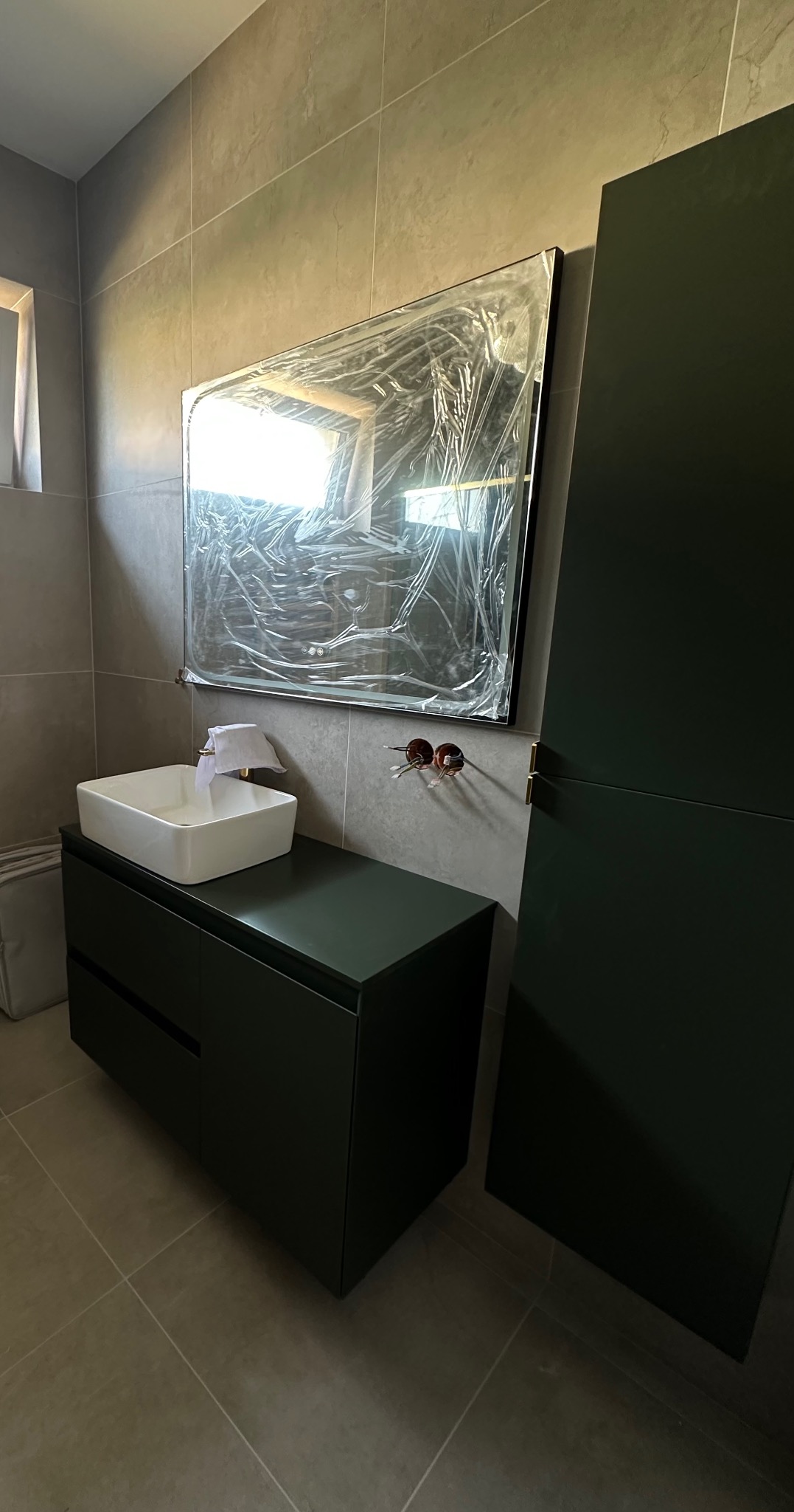 Nowoczesna łazienka w trakcie wykańczania: ciemnozielona szafka z umywalką, lustro w folii ochronnej, widoczne instalacje elektryczne i płytki ceramiczne.