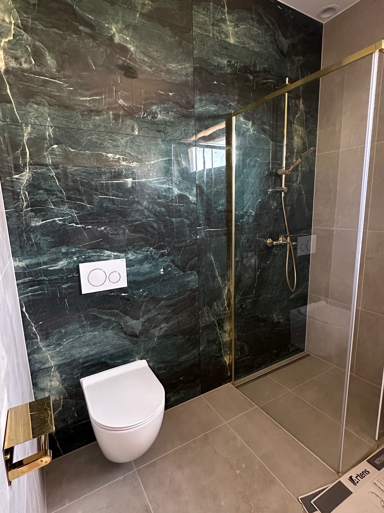 Nowoczesna łazienka z kabiną prysznicową w złotych ramach i toaletą podwieszaną na tle ściany z zielonego marmuru. Widoczne szare płytki podłogowe.