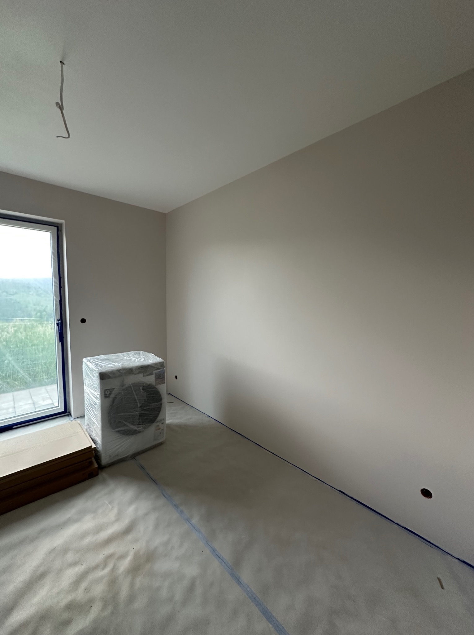 Pokój po malowaniu ścian na szaro, zabezpieczony folią i taśmą malarską. Okno z widokiem na zieleń. Nowa pralka w folii ochronnej w rogu pokoju.