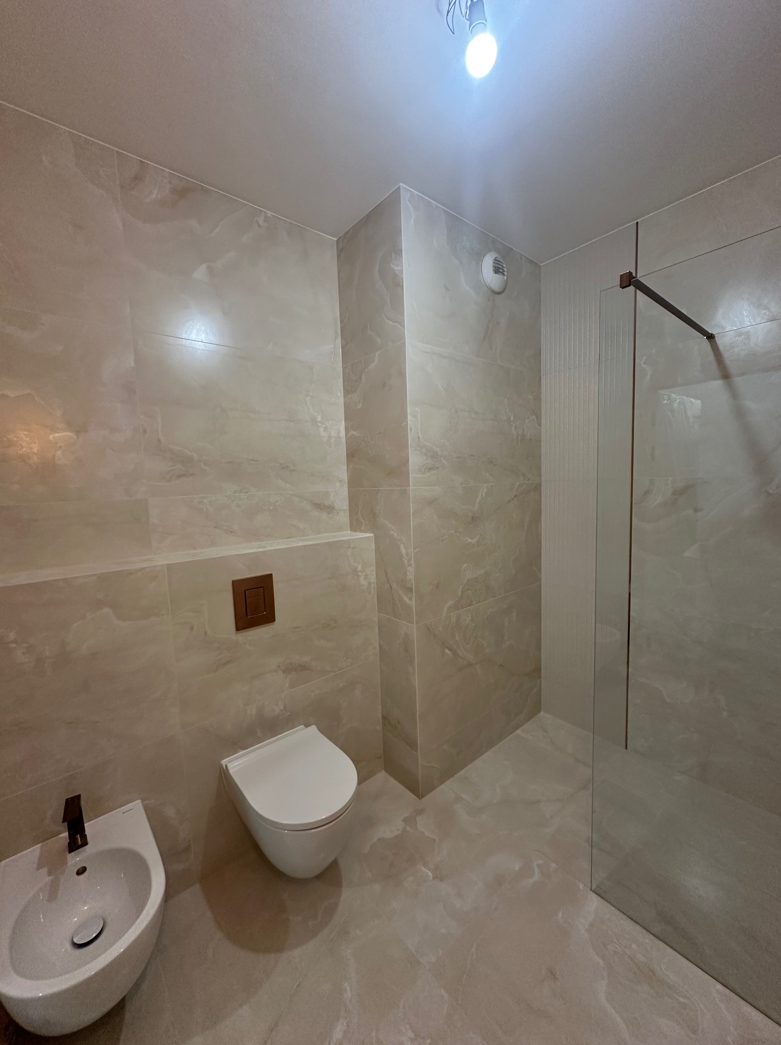 Wykończona łazienka z beżowymi płytkami imitującymi marmur, wisząca toaleta z brązowym przyciskiem, bidet z czarną armaturą i szklana kabina prysznicowa.