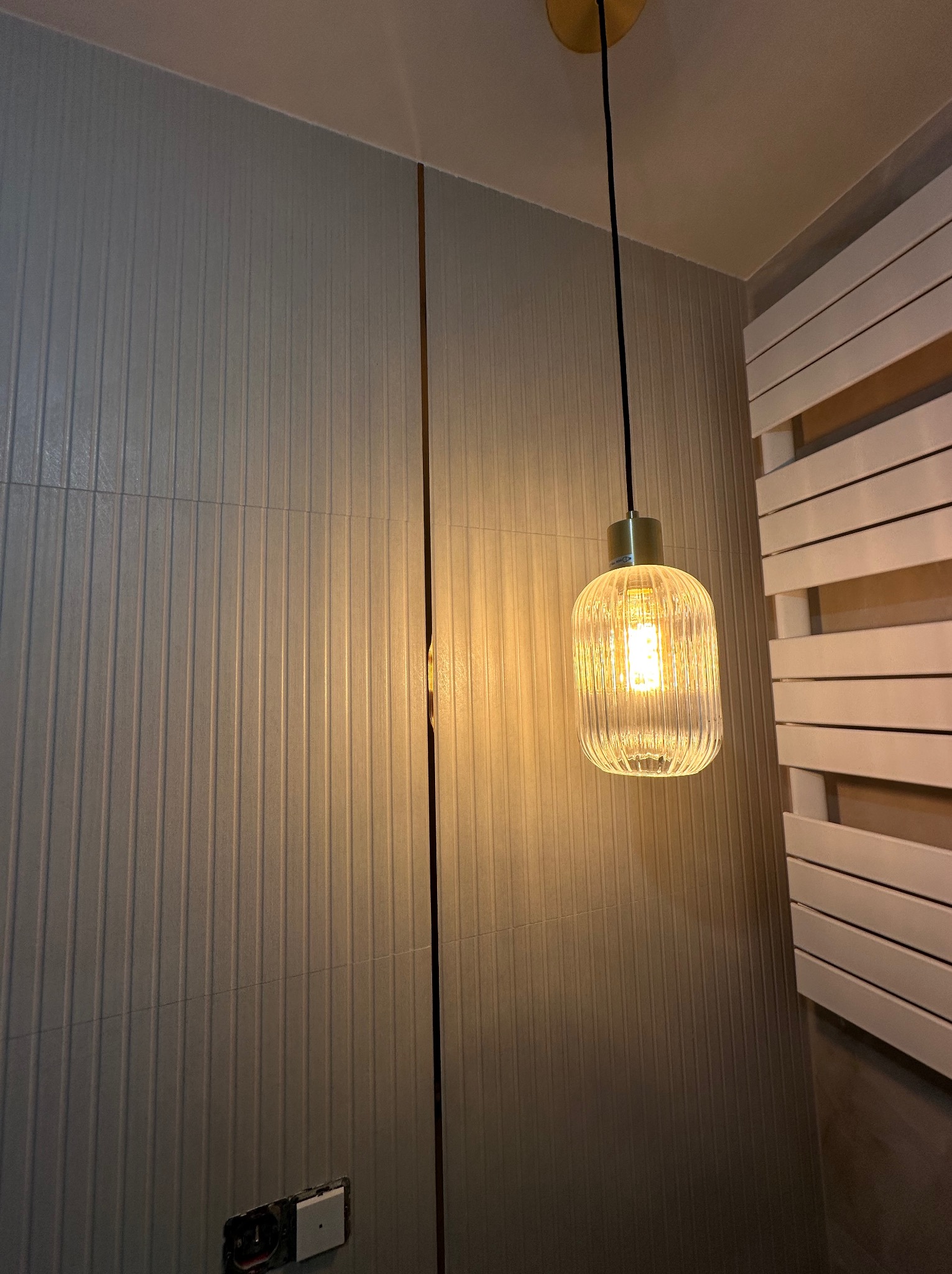 Nowoczesna łazienka z szarymi płytkami ściennymi, dekoracyjnym grzejnikiem drabinkowym i wiszącą lampą z żarówką o ciepłej barwie.