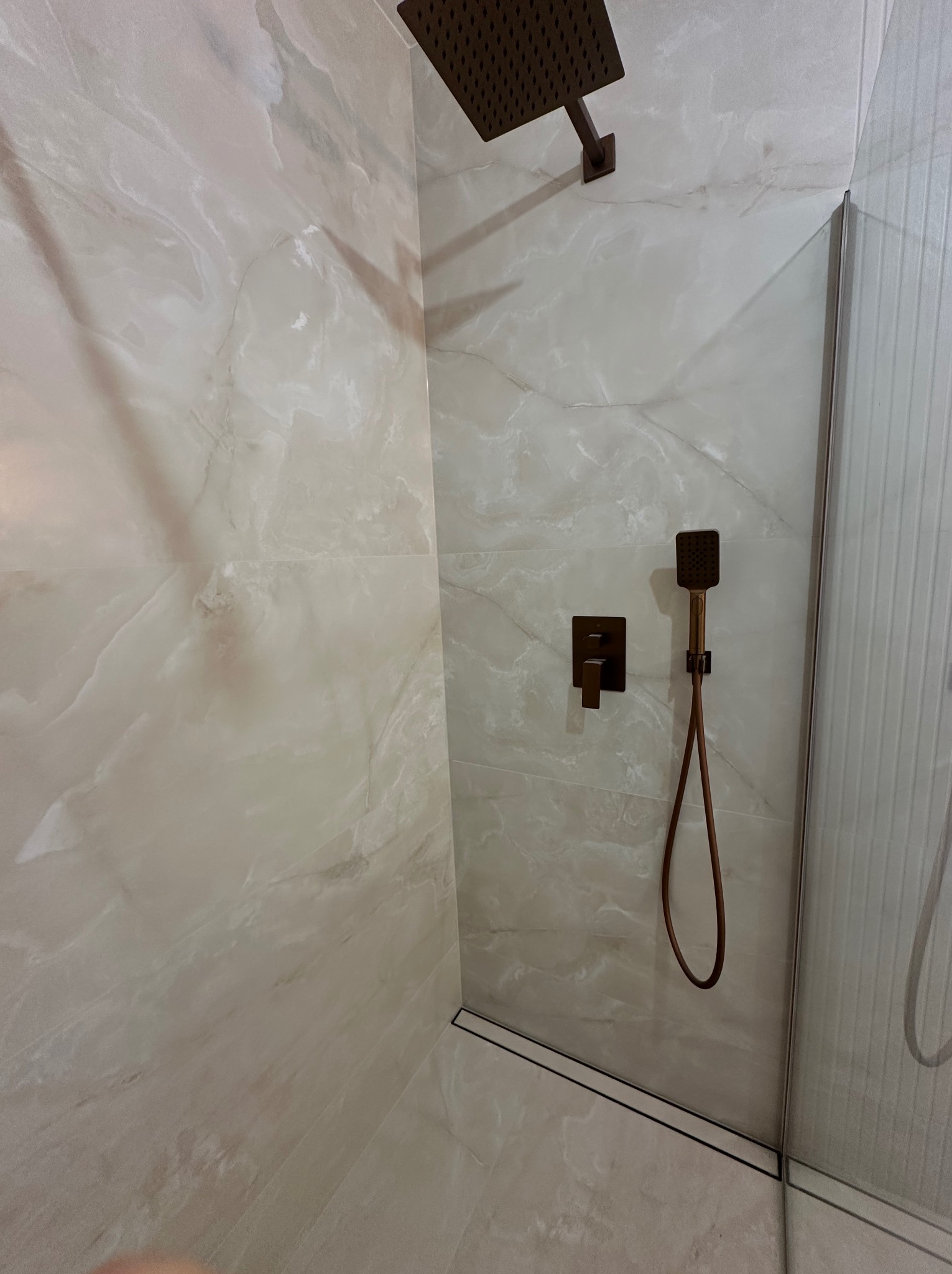 Nowoczesna kabina prysznicowa z marmurowymi płytkami, deszczownicą i baterią w kolorze brązu. Minimalistyczny design z odpływem liniowym i szklaną ścianką.