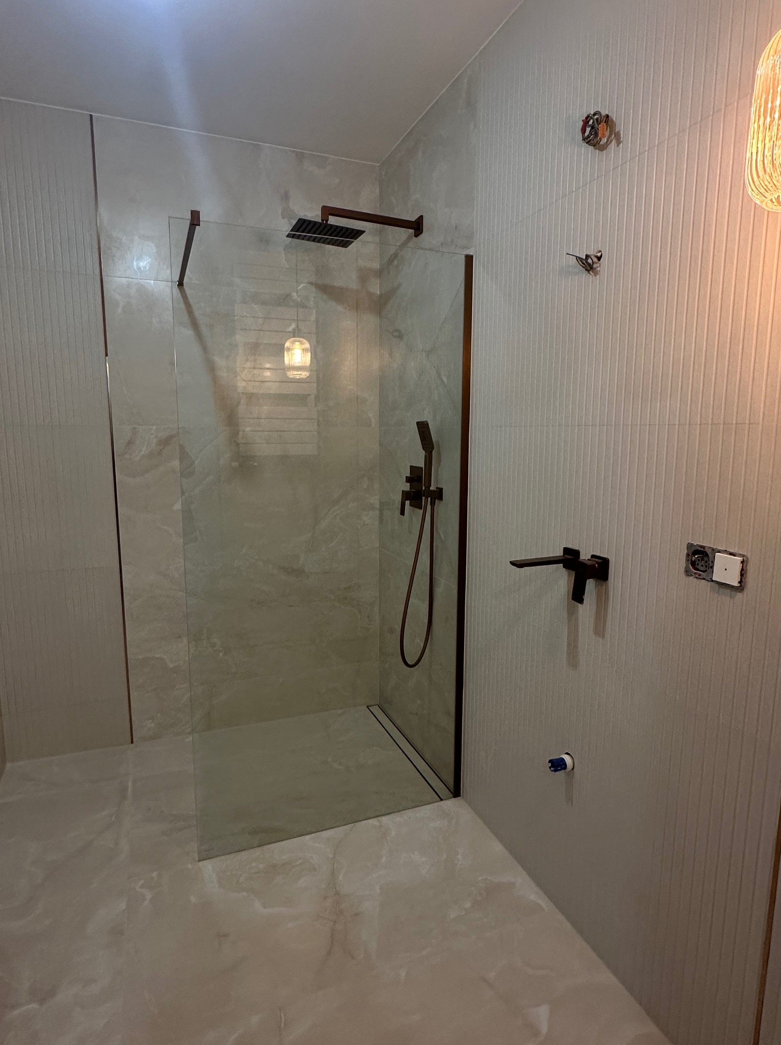 Nowoczesna kabina prysznicowa walk-in z czarną armaturą i szklaną ścianką, wykończona płytkami w odcieniach beżu i szarości. Widoczne detale instalacji.