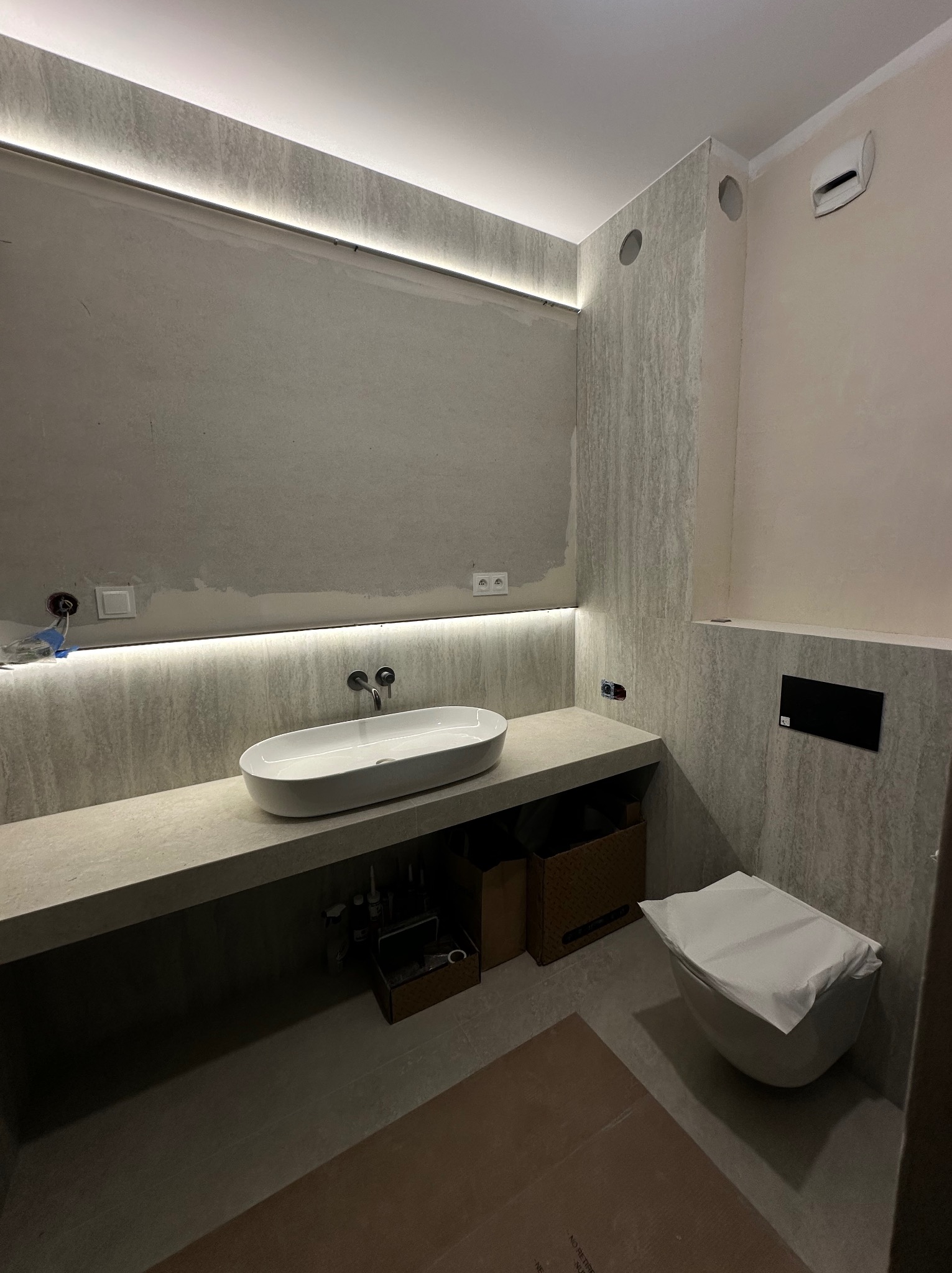 Nowoczesna łazienka w trakcie wykończenia: owalna umywalka na blacie z podświetleniem LED, toaleta z zabezpieczeniem, szare płytki imitujące kamień.