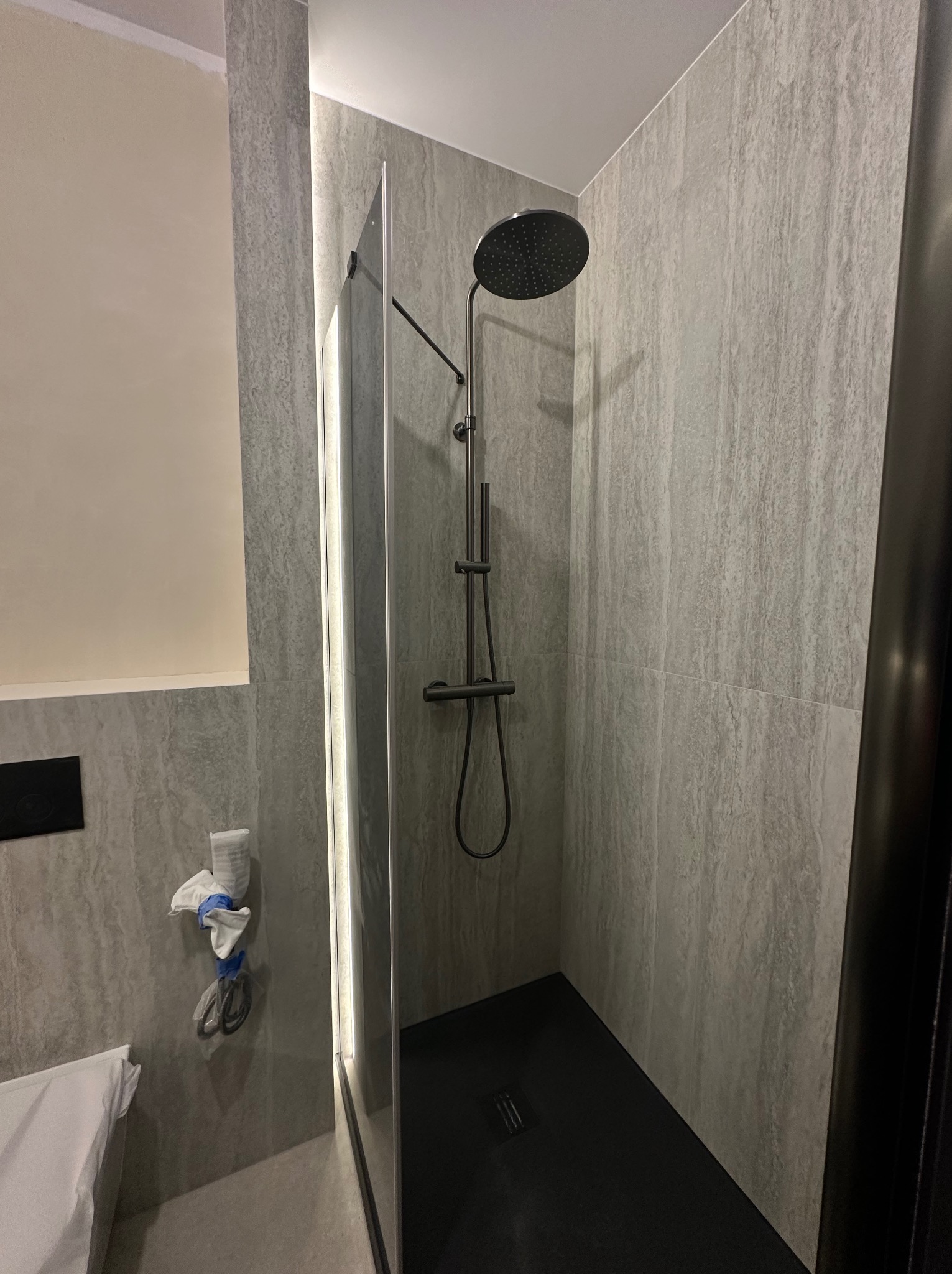 Nowoczesna kabina prysznicowa z deszczownicą i szklaną ścianką, wykończona szarymi płytkami imitującymi kamień. Widoczne akcesoria łazienkowe i czarna podłoga.