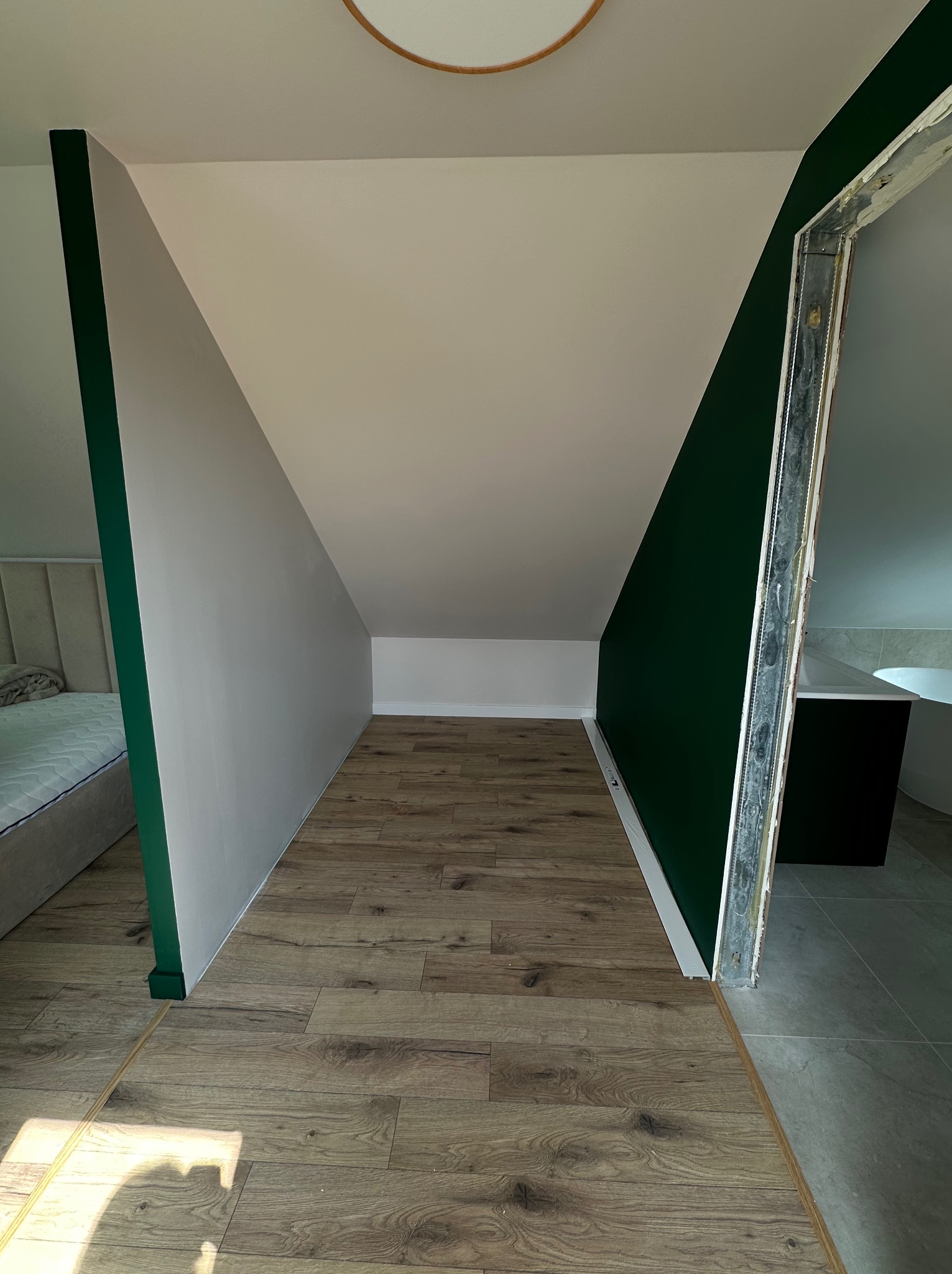 Wykończone poddasze ze skośnymi ścianami pomalowanymi na biało i butelkową zieleń. Widoczna podłoga z paneli i fragment łazienki z wanną. Metalowa futryna.