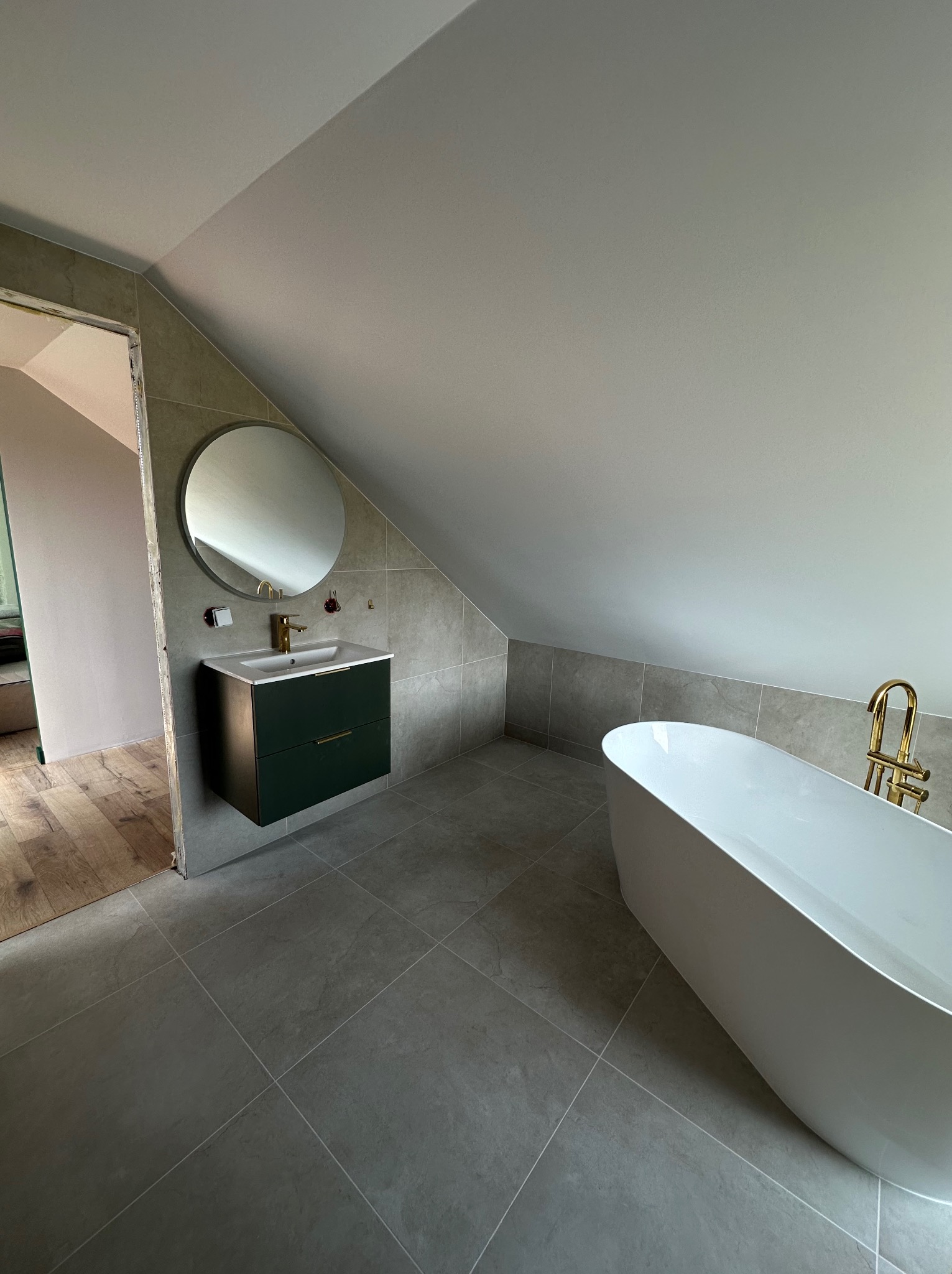 Nowoczesna łazienka na poddaszu z wolnostojącą wanną, ciemnozieloną szafką z okrągłym lustrem i złotą armaturą. Szare płytki na ścianach i podłodze.