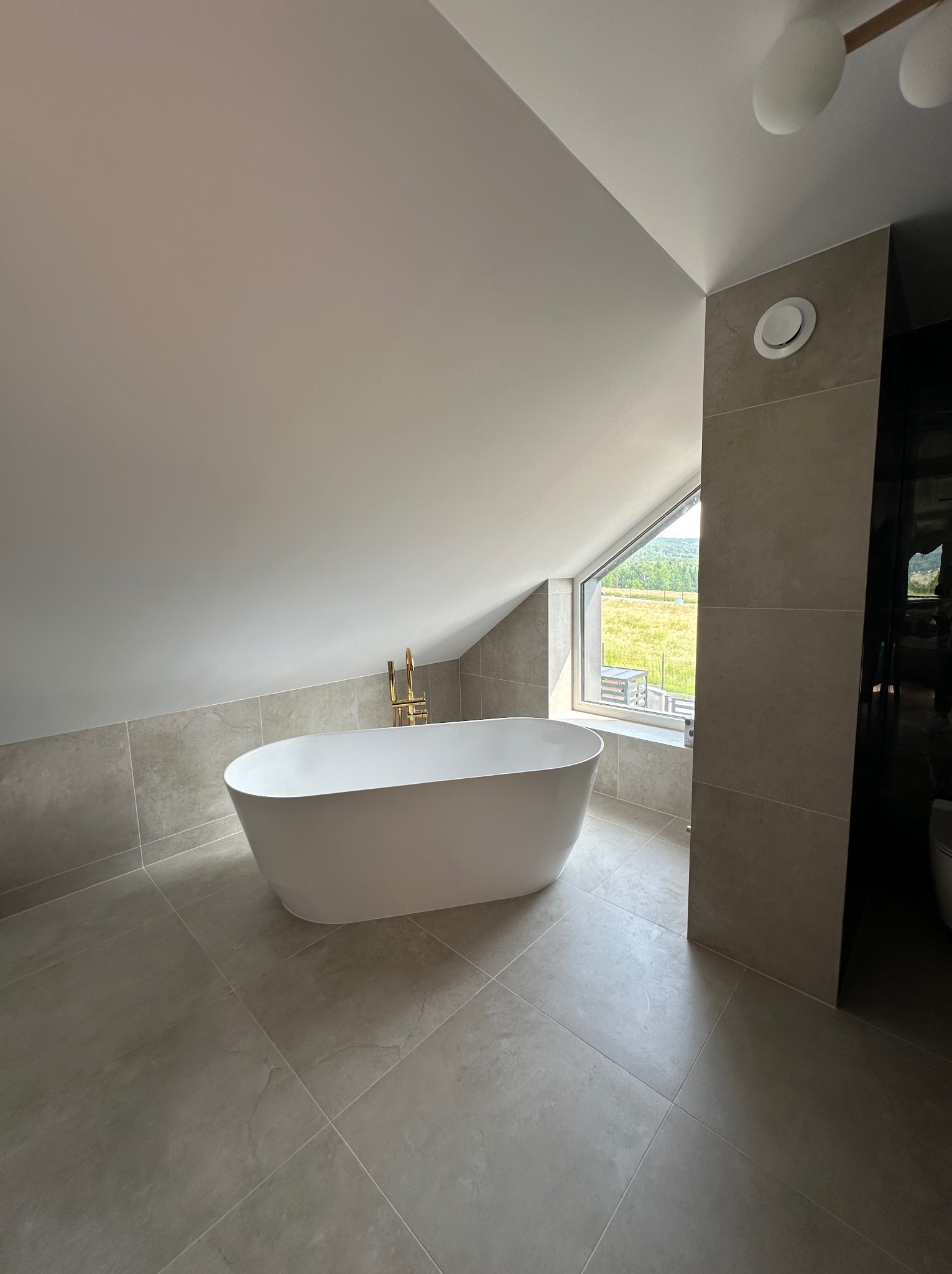 Nowoczesna, jasna łazienka na poddaszu z wolnostojącą wanną, złotą armaturą i oknem z widokiem na zieleń. Minimalistyczny design z szarymi płytkami i subtelnym oświetleniem.