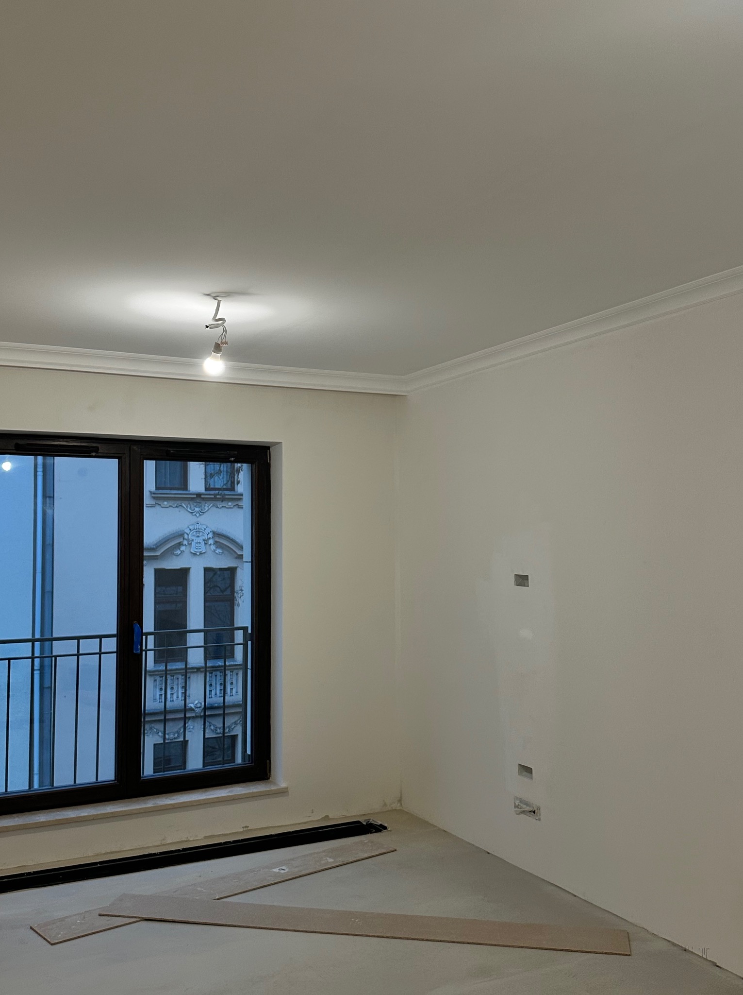 Jasne wnętrze w trakcie prac wykończeniowych: białe ściany z listwami, okno balkonowe z widokiem na budynek, fragmenty paneli podłogowych na podłodze.