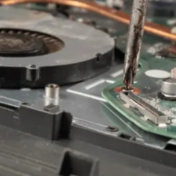 Otwarty laptop, widoczna płyta główna i wentylator, śrubokręt wkręca śrubkę przy złączu.