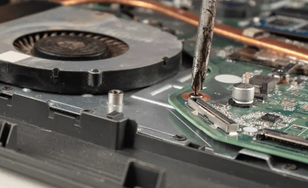 Otwarty laptop, widoczna płyta główna i wentylator, śrubokręt wkręca śrubkę przy złączu.