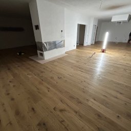 WOODEAL SPÓŁKA Z OGRANICZONĄ ODPOWIEDZIALNOŚCIĄ - Nowo ułożona, jasna, drewniana podłoga w przestronnym wnętrzu z kominkiem i oświetleniem roboczym, widoczne ślady po pracach wykończeniowych.