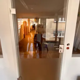 Szklane drzwi wewnętrzne z białą ramą, widok na korytarz z parkietem i balkonem w tle.