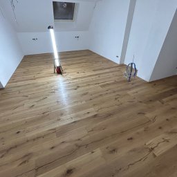 WOODEAL SPÓŁKA Z OGRANICZONĄ ODPOWIEDZIALNOŚCIĄ - Ukończona podłoga z paneli drewnianych w jasnym pokoju na poddaszu, oświetlona lampą budowlaną. Widoczne gniazdka elektryczne i kable.