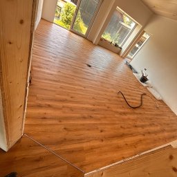 WOODEAL SPÓŁKA Z OGRANICZONĄ ODPOWIEDZIALNOŚCIĄ - Ukończone układanie paneli podłogowych w jasnym wnętrzu z oknami. Widoczne detale łączeń i wykończenia przy ścianach. Częściowo widoczne narzędzia i materiały.