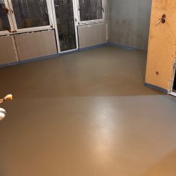 WOODEAL SPÓŁKA Z OGRANICZONĄ ODPOWIEDZIALNOŚCIĄ - Świeżo wylana, gładka posadzka jastrychowa w pomieszczeniu z oknami i przygotowanymi ścianami do malowania. Widoczne zabezpieczenia folią przy ścianach.