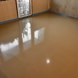 WOODEAL SPÓŁKA Z OGRANICZONĄ ODPOWIEDZIALNOŚCIĄ - Świeżo wylana, brązowa posadzka jastrychowa w pomieszczeniu z oknem. Widoczne ściany w stanie surowym i listwa dylatacyjna przy ścianie. Gładka, błyszcząca powierzchnia.