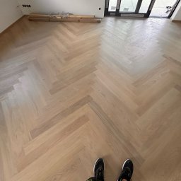 WOODEAL SPÓŁKA Z OGRANICZONĄ ODPOWIEDZIALNOŚCIĄ - Jasny parkiet ułożony we wzór jodełki francuskiej, widok z góry na fragment podłogi z parą butów w dolnej części kadru, w tle deski do ułożenia.