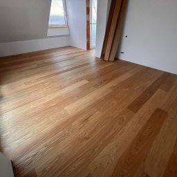 WOODEAL SPÓŁKA Z OGRANICZONĄ ODPOWIEDZIALNOŚCIĄ - Ukończona podłoga z paneli w jasnym odcieniu drewna w pokoju na poddaszu. Widoczne deski podłogowe i fragment okna dachowego. Częściowo zakryte folią zabezpieczającą.