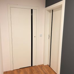 WOODEAL SPÓŁKA Z OGRANICZONĄ ODPOWIEDZIALNOŚCIĄ - Dwa białe, nowoczesne drzwi wewnętrzne z czarnymi akcentami na tle drewnopodobnej podłogi i ściany w odcieniach bieli i grafitu. Widoczny fragment włącznika światła.