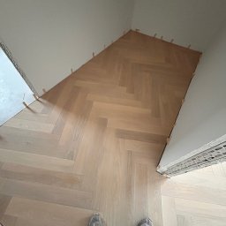 WOODEAL SPÓŁKA Z OGRANICZONĄ ODPOWIEDZIALNOŚCIĄ - Jasny parkiet ułożony we wzór jodełki, z widocznymi klinami dystansowymi przy ścianach. U dołu kadru stopy w roboczych butach. Ujęcie z góry, w trakcie prac.