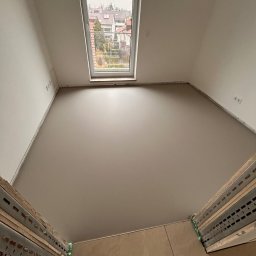 WOODEAL SPÓŁKA Z OGRANICZONĄ ODPOWIEDZIALNOŚCIĄ - Poddasze po gładziowaniu ścian i sufitu, widok na okno dachowe i fragment podłogi. Metalowe profile w dolnej części kadru. Świeżo wykończone wnętrze.