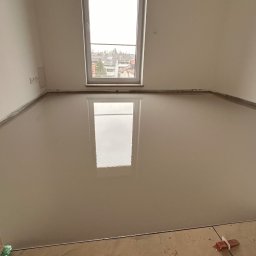 WOODEAL SPÓŁKA Z OGRANICZONĄ ODPOWIEDZIALNOŚCIĄ - Świeżo wylana, gładka posadzka anhydrytowa w jasnym pomieszczeniu z oknem, odbijającym światło. Widoczne listwy przypodłogowe i fragment rusztowania.