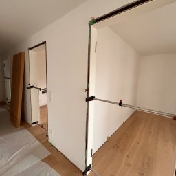 WOODEAL SPÓŁKA Z OGRANICZONĄ ODPOWIEDZIALNOŚCIĄ - Wnętrze z zamontowanymi ościeżnicami drzwiowymi, zabezpieczonymi rozpórkami. Na podłodze widoczny parkiet, częściowo zakryty folią ochronną. Ściany pomalowane na biało.