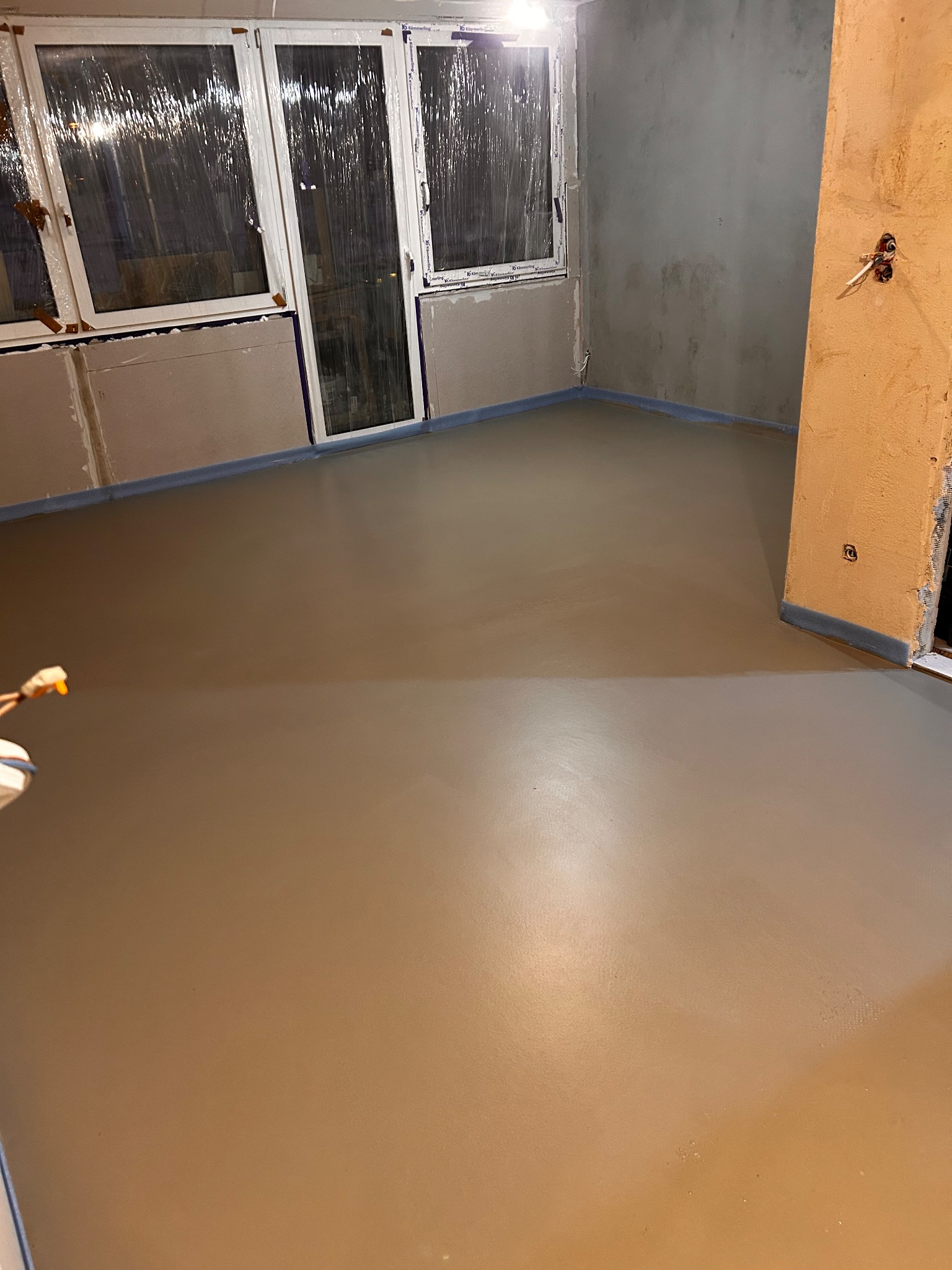 Świeżo wylana, gładka posadzka jastrychowa w pomieszczeniu z oknami i przygotowanymi ścianami do malowania. Widoczne zabezpieczenia folią przy ścianach.